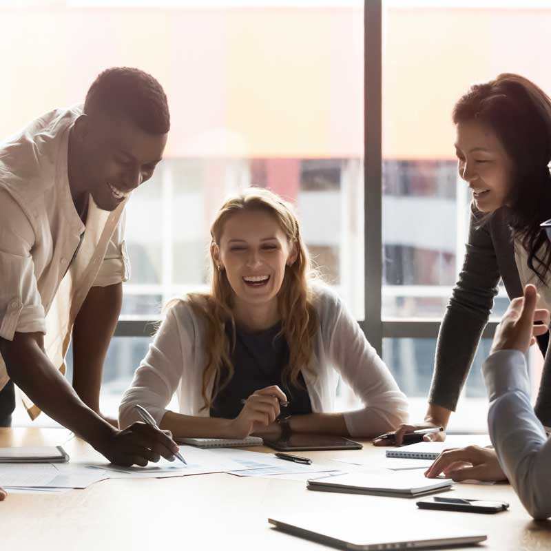 Smiling colleagues gather around a table with papers and tablets, engaged in a lively discussion. Bright window light creates a warm, collaborative atmosphere.