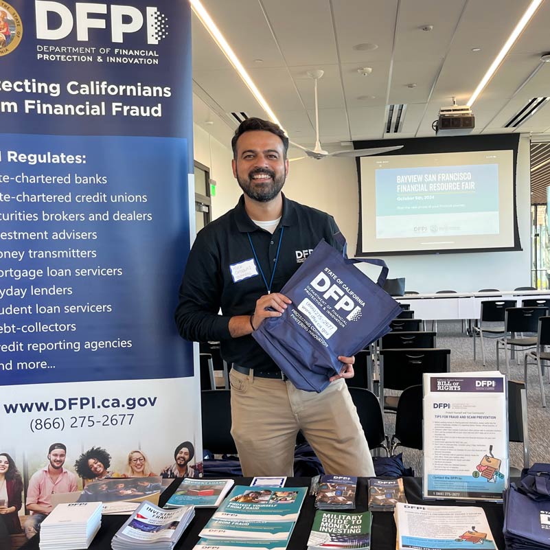Smiling man at a financial resource fair holds a DFPI tote bag. A banner displays fraud prevention resources. Flyers and brochures are neatly arranged on a table.